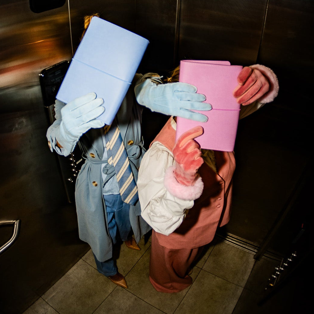 Two people standing inside a metallic elevator holding colorful folders, one with a light blue folder and the other with a pink folder, wearing oversized pastel-colored face masks and gloves.